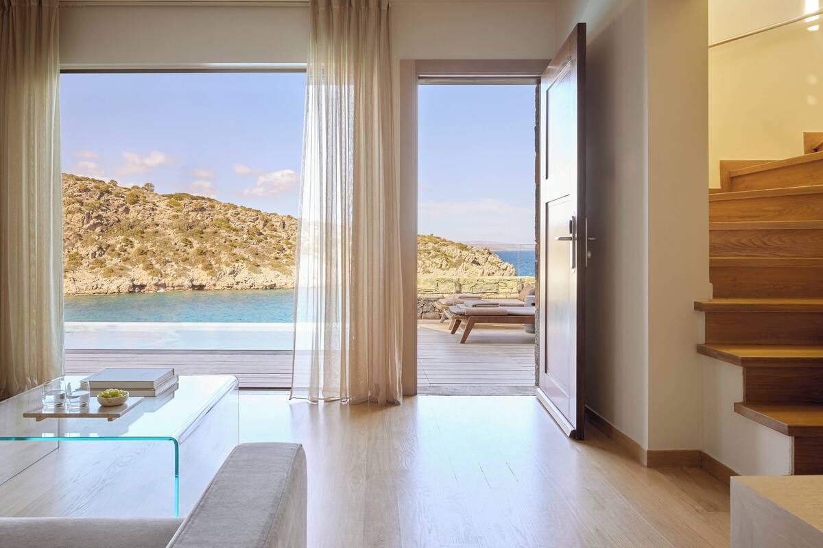 A modern room with a glass table, a view of the sea, and a wooden staircase.
