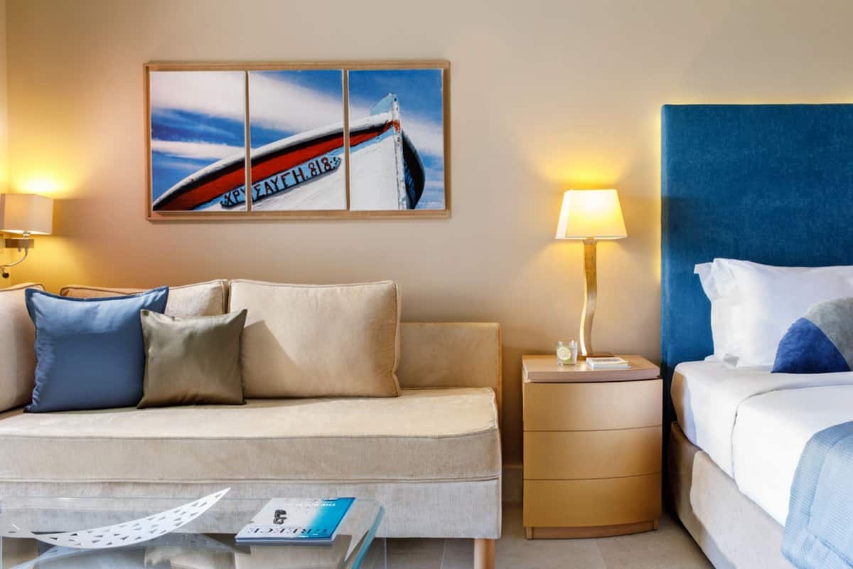 A hotel room with a sofa, a bed, a nightstand, a lamp, and a painting on the wall.