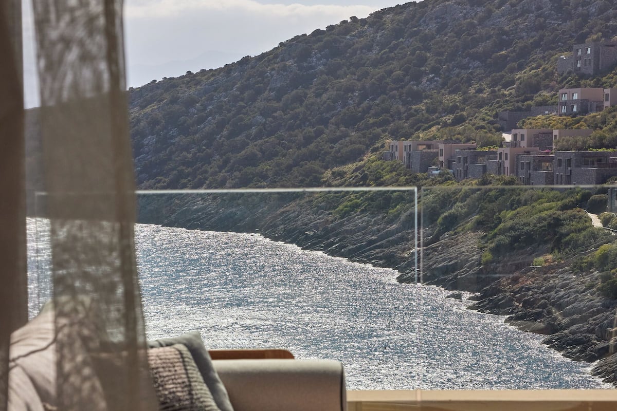 A modern outdoor seating area with a sofa and a round table, overlooking a beach and a hillside with buildings.