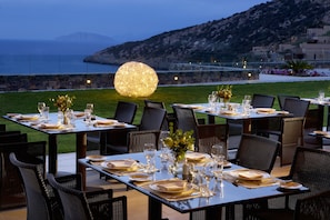 An outdoor dining area with tables set for a meal, overlooking a body of water and hills. - Daios Cove (Agios Nikolaos)