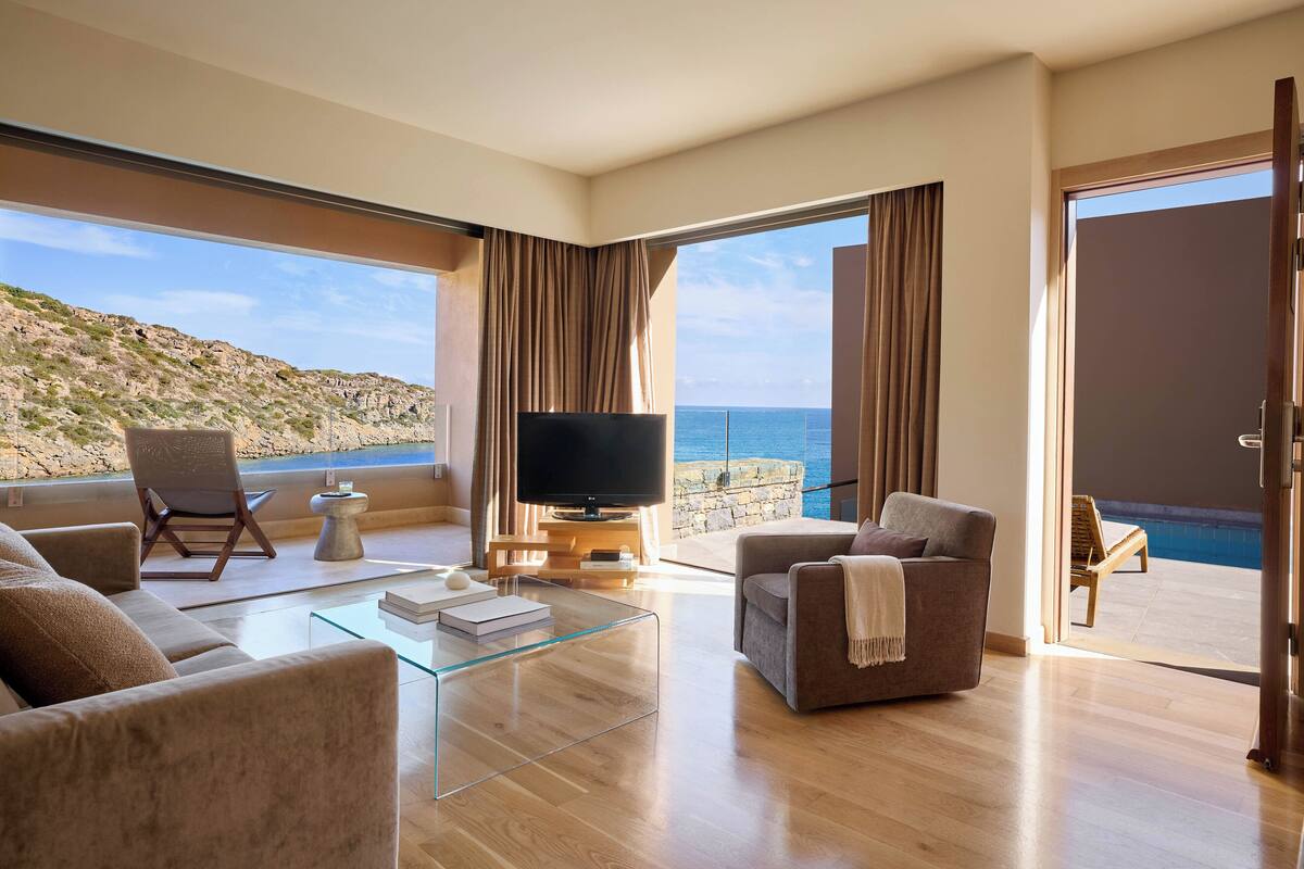A modern living room with a large TV, comfortable seating, a glass coffee table, and a view of the sea.