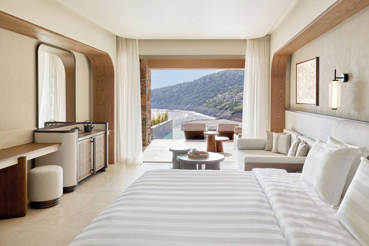 A modern hotel room with a large bed, a sitting area with a sofa and chairs, a small table, and a view of a mountainous landscape through a large window.