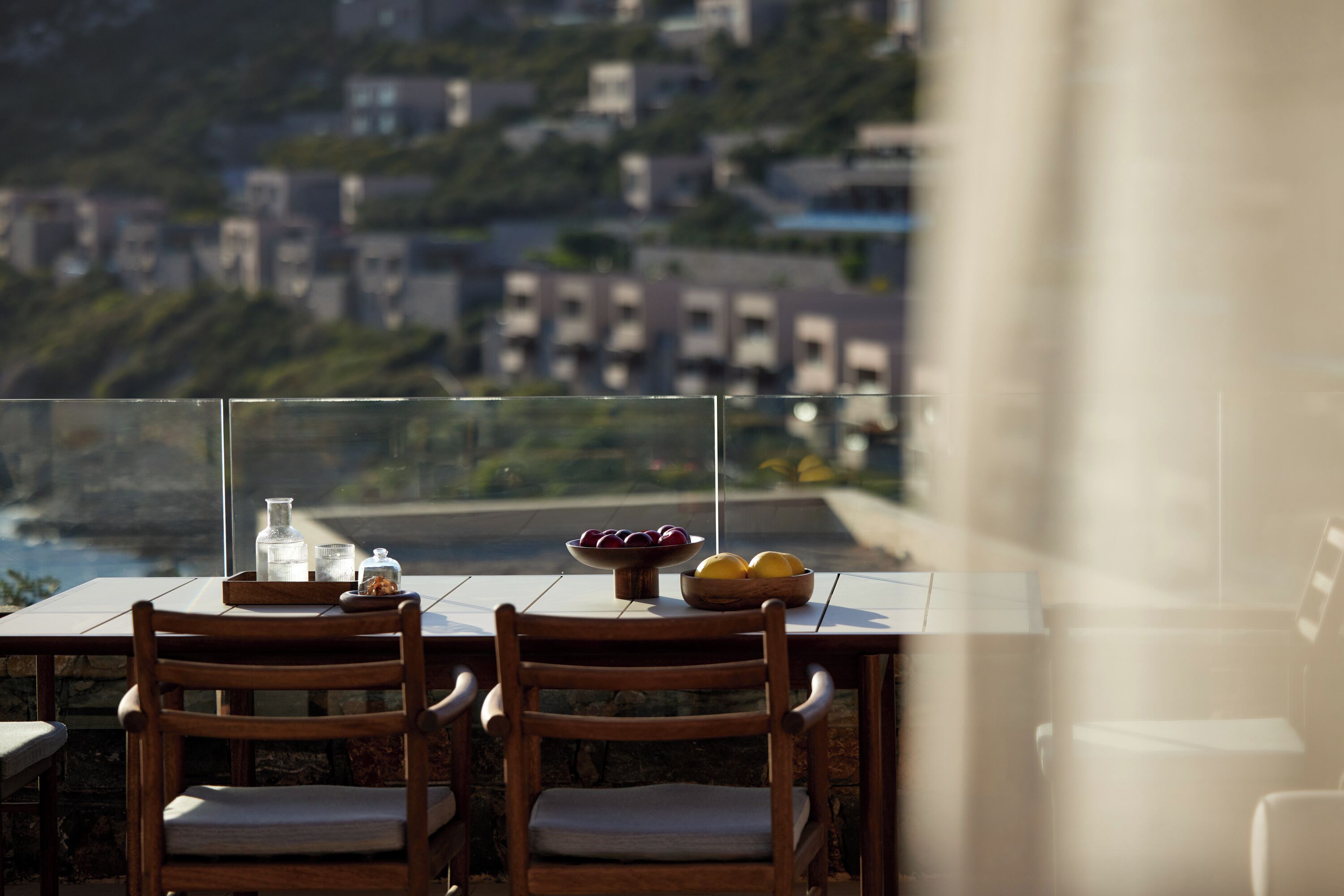 Ein Esstisch mit Stühlen, eine Obstschale und ein Glasgefäß auf einem Balkon mit Blick auf eine Küstenlandschaft und Gebäude.