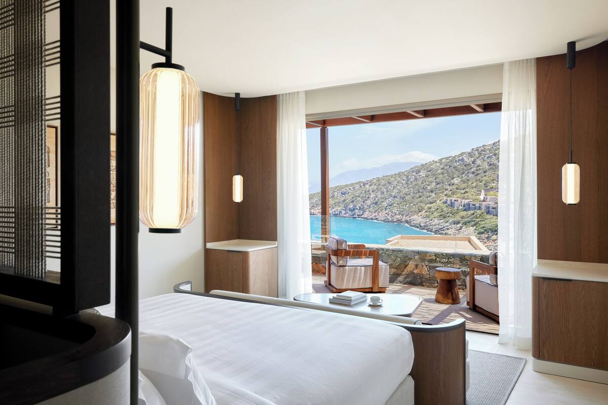 A modern hotel room with a large bed, a seating area with a view of the sea, and a balcony with a pool.