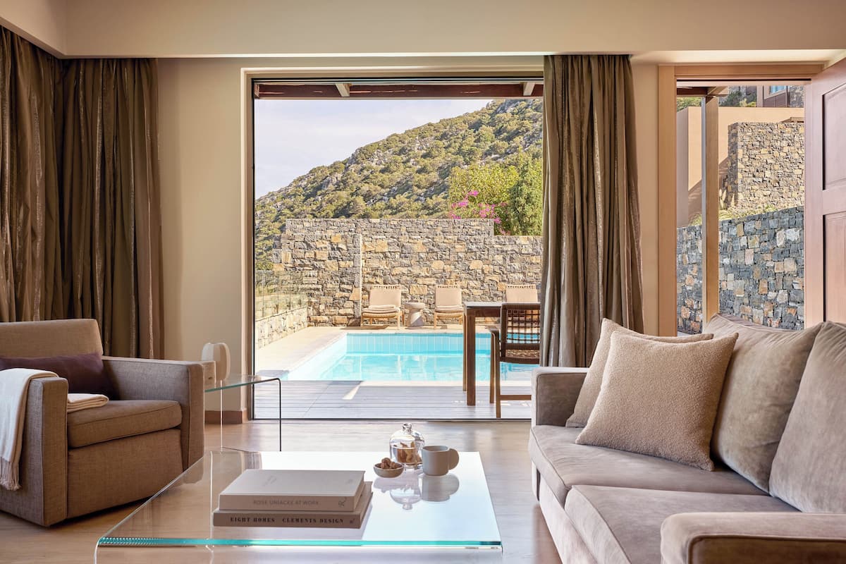 A modern living room with a glass coffee table, a sofa, and a view of a pool and stone wall.