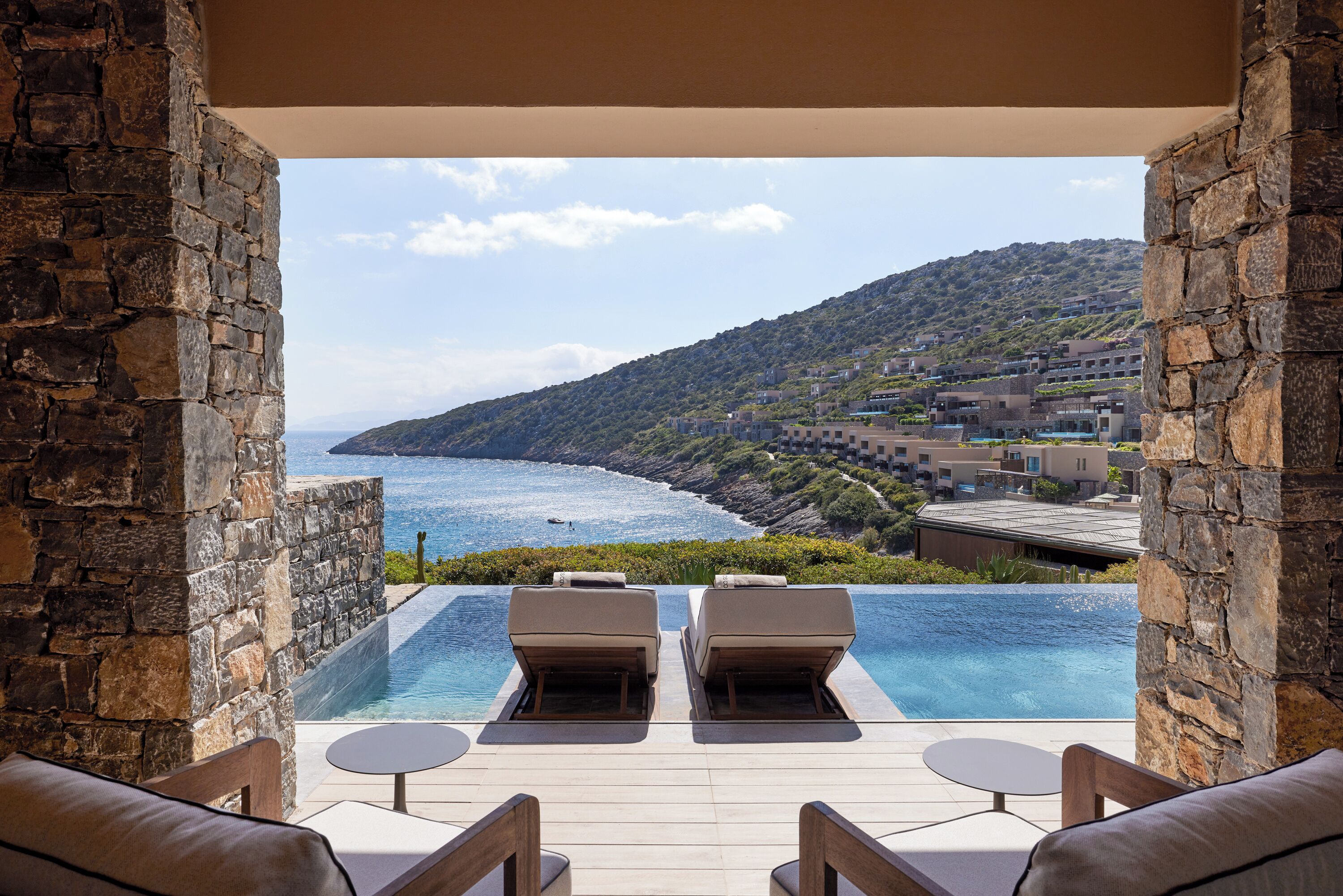An infinity pool with lounge chairs, stone walls, and a view of the sea and hills.