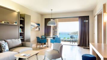 Un salon moderne avec un canapé, un fauteuil et une table basse. On y trouve également un coin repas avec une table et des chaises, et une vue sur la mer grâce à de grandes fenêtres.