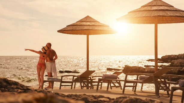 A beach scene with two thatched umbrellas and lounge chairs, a couple standing by the water, and a rocky shoreline. - Valamar Riviera Hotel & Residence (Porec)