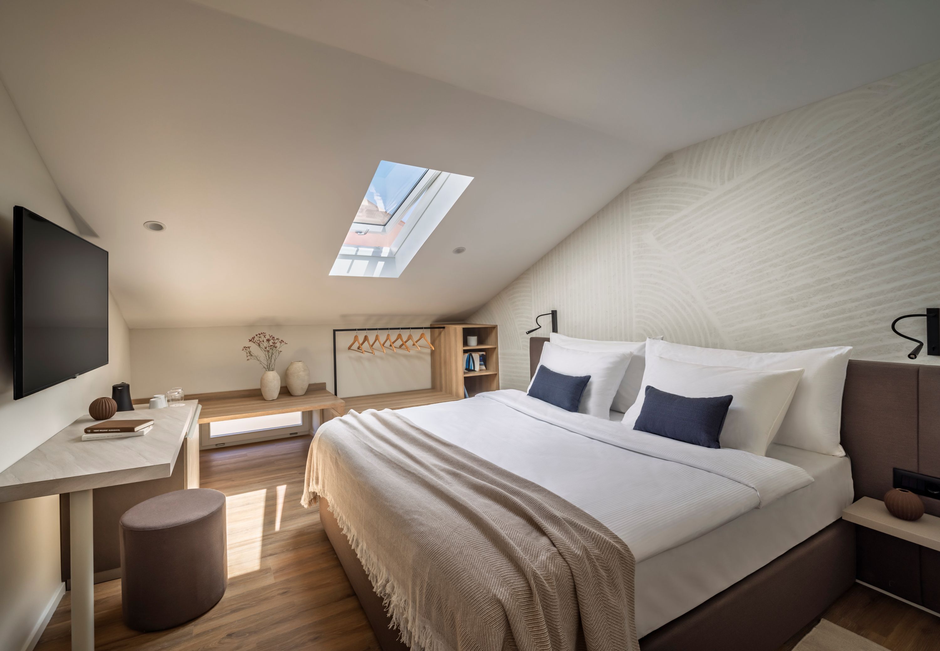 A modern bedroom with a sloped ceiling, a large bed with white and blue bedding, a desk with a chair, a small stool, and a TV mounted on the wall.