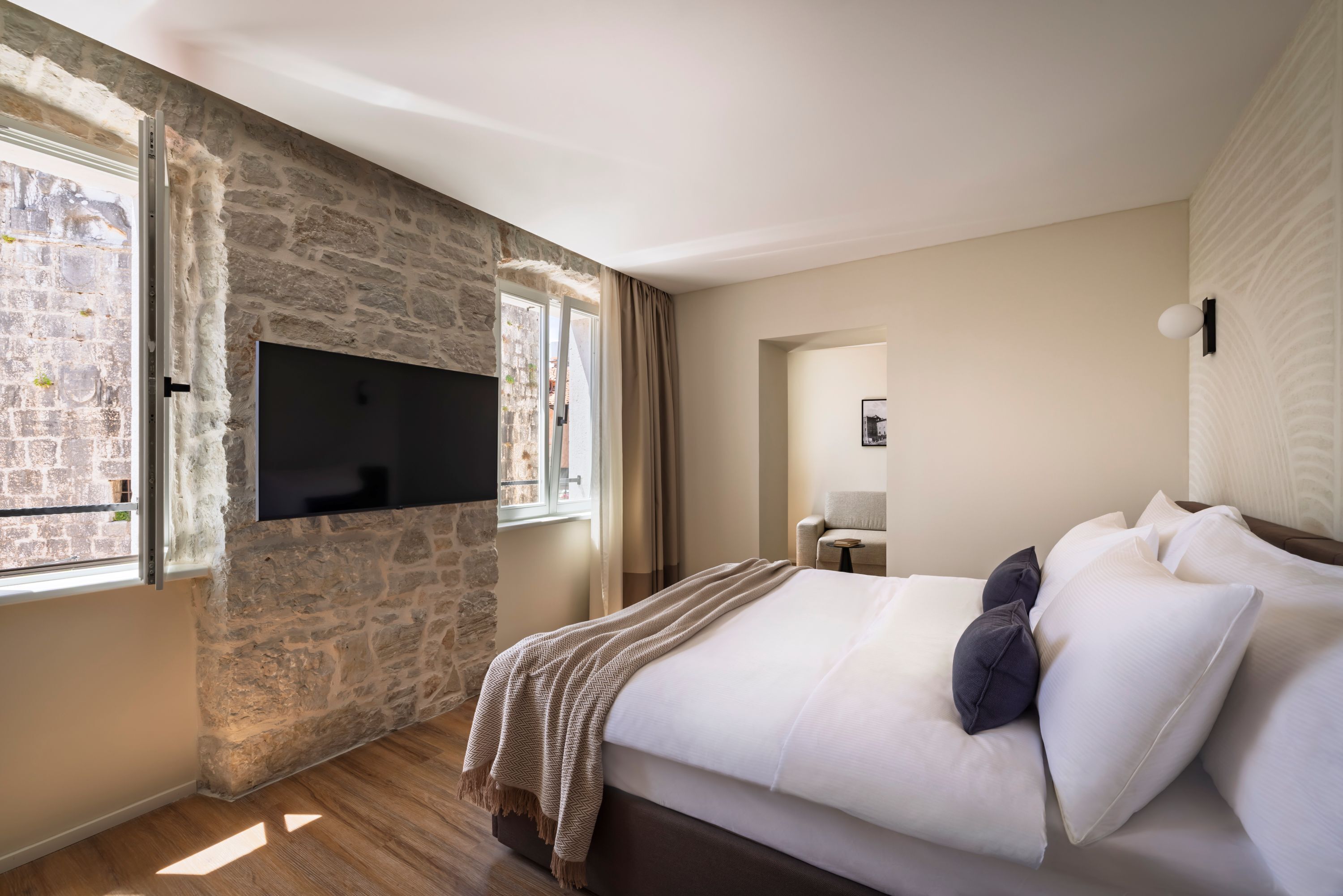 A bedroom with a stone wall, a flat-screen TV, a bed with white and dark pillows, and a view through a window.