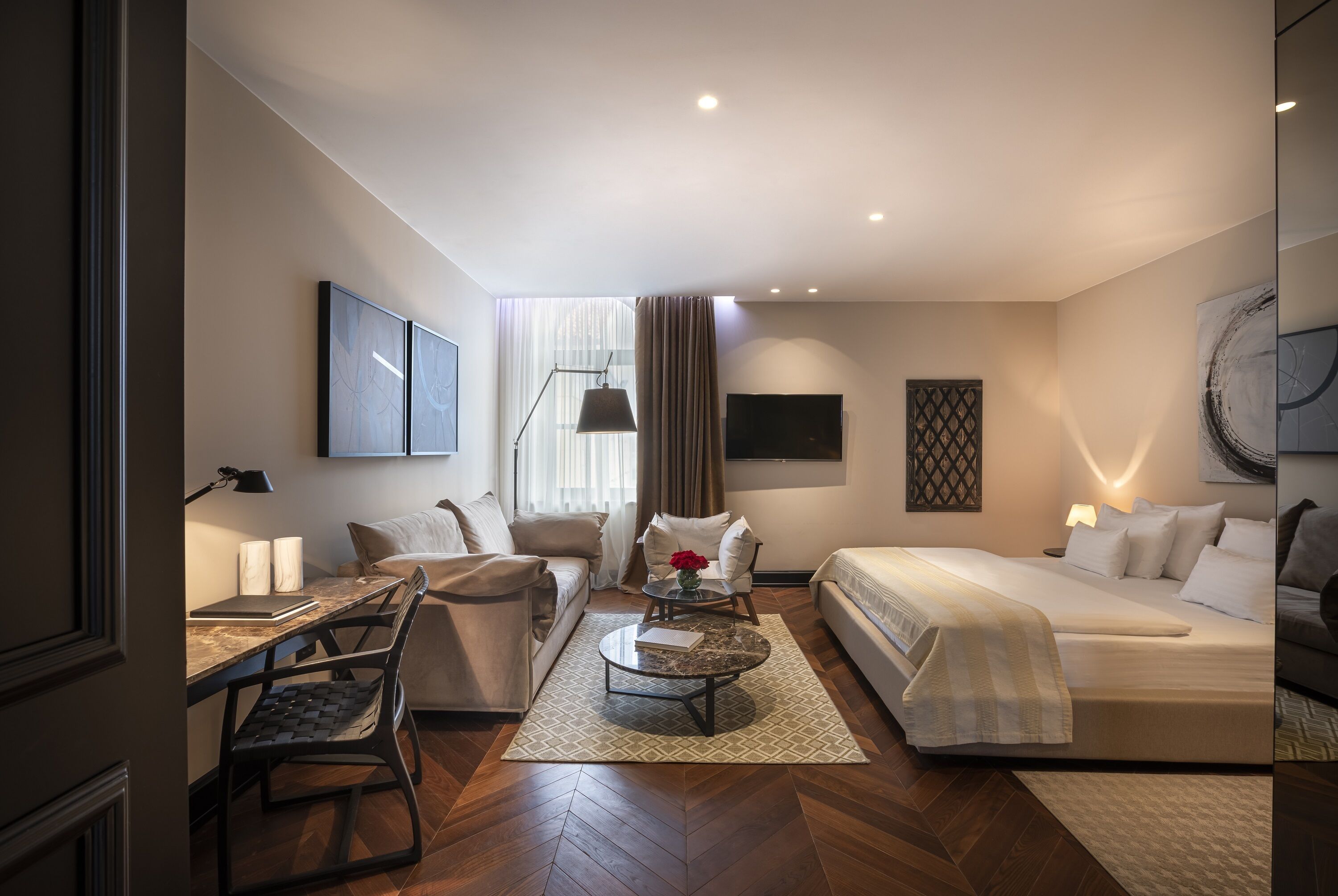 A modern hotel room with a sofa, a bed, a desk with a lamp, and a coffee table.