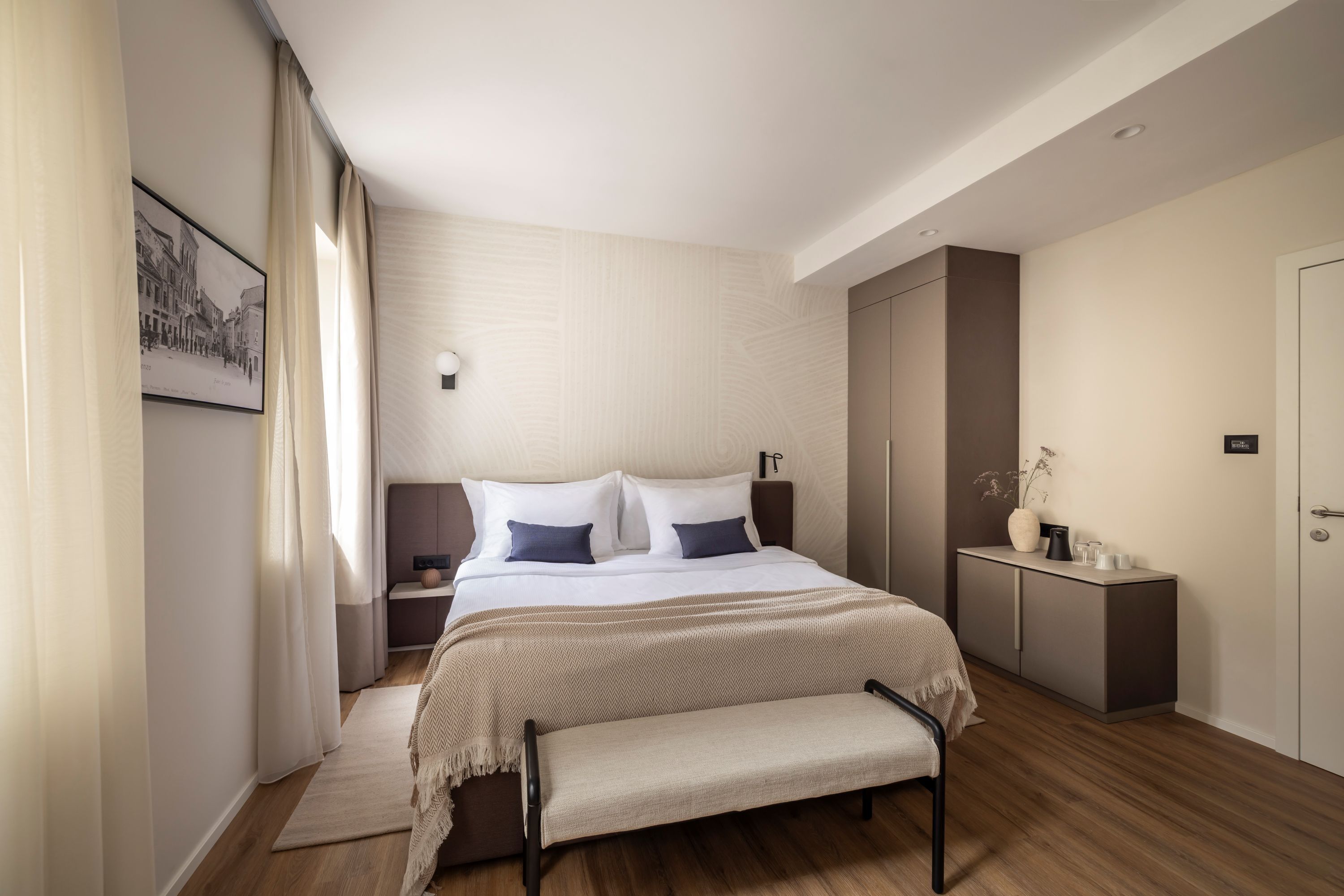 A modern bedroom with a large bed, bedside tables, a bench, a wardrobe, and a framed picture on the wall.