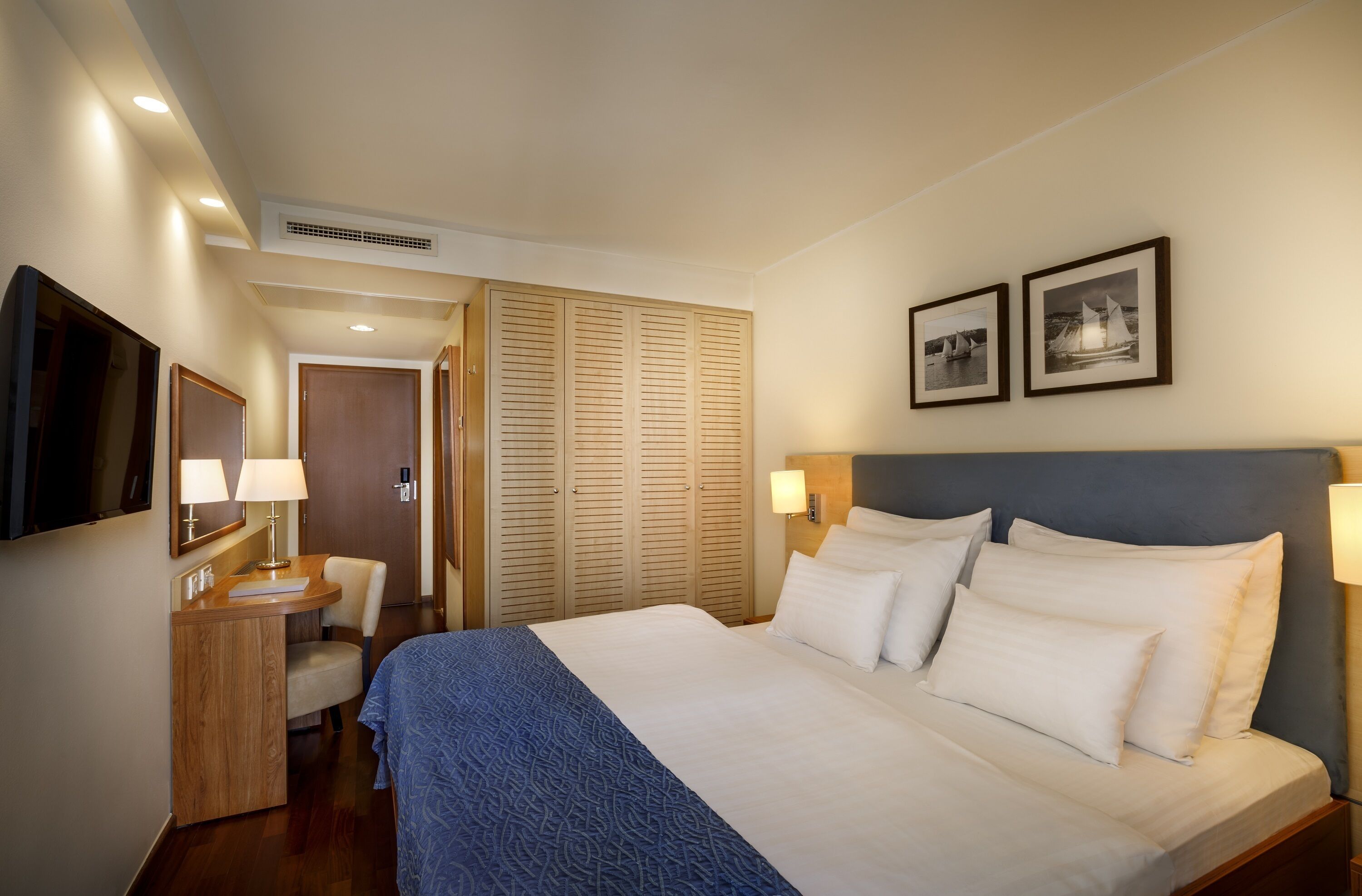Hotelkamer met een bed, bedlampen, een bureau met stoel, een kledingkast en twee ingelijste foto's aan de muur.