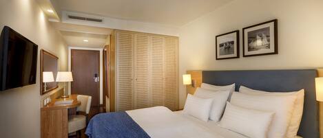 Une chambre d’hôtel comprenant un lit, des lampes de chevet, un bureau avec une chaise, une armoire et deux tableaux encadrés accrochés au mur.