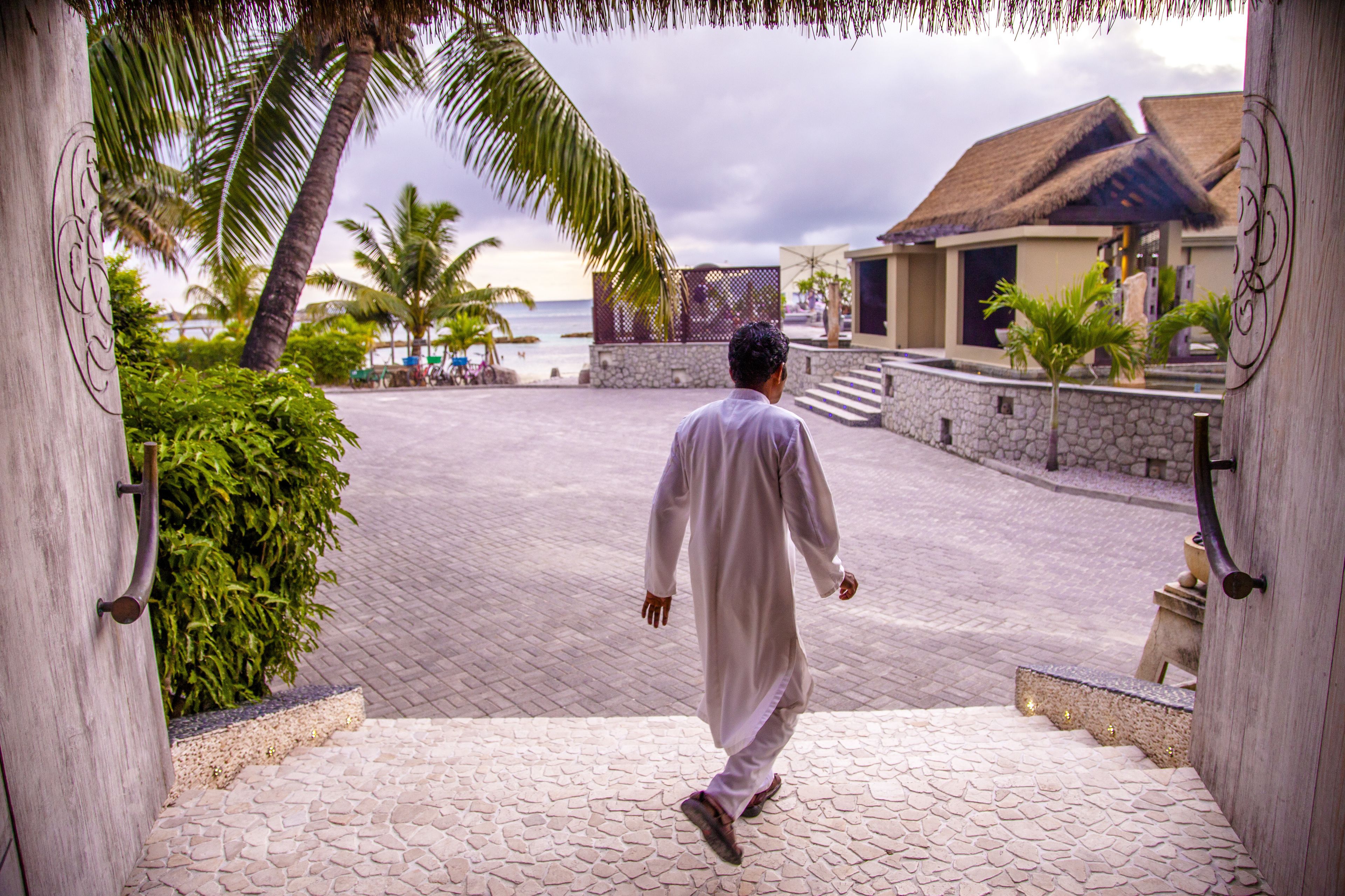 A man walking through a paved area with palm trees and a building with a thatched roof in the background.
