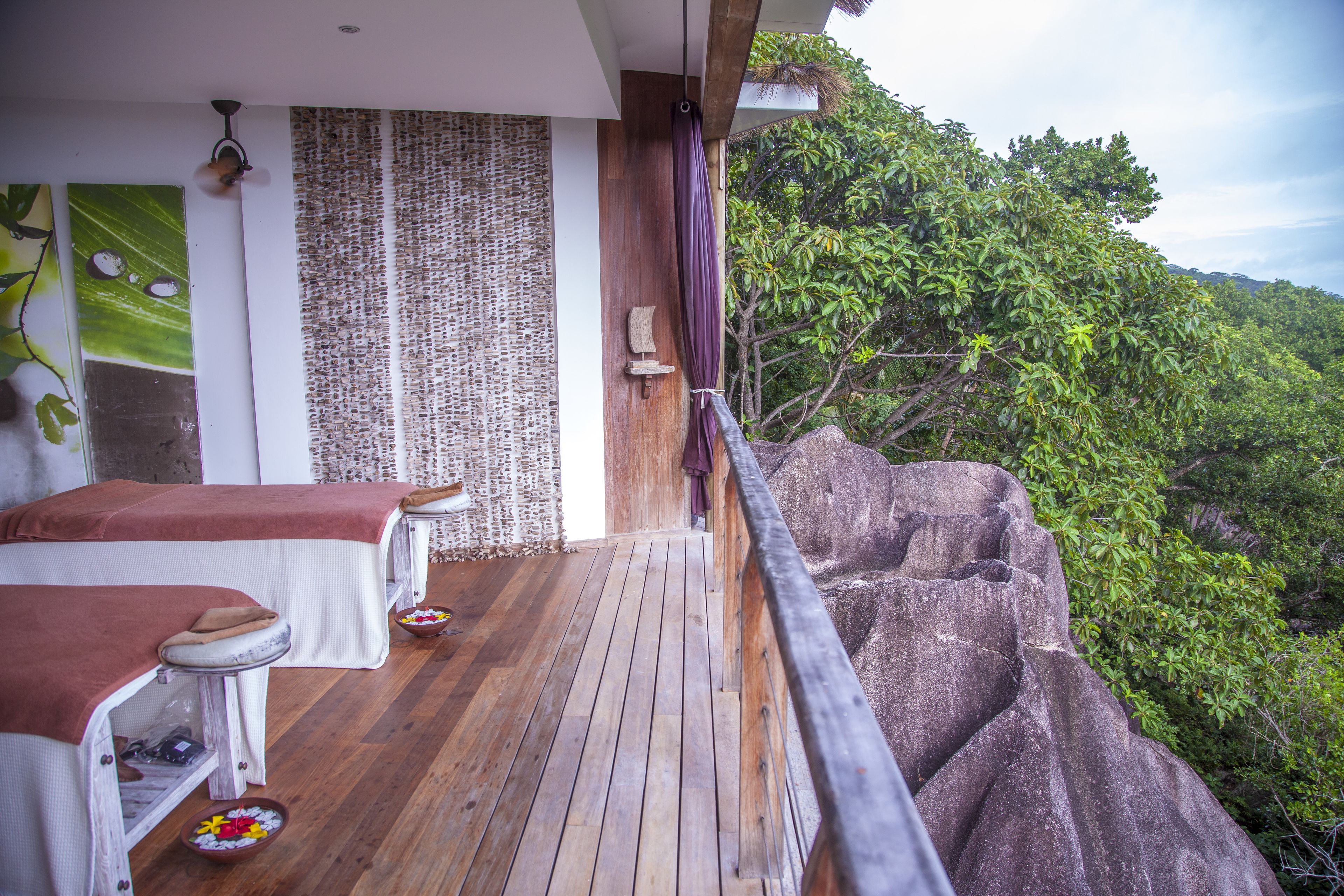 A wooden deck with massage tables, a woven wall, and a view of lush greenery.