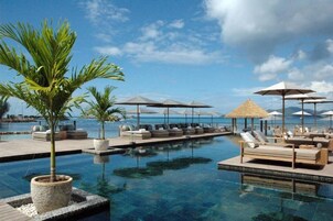 Zona junto a la piscina con sillones de descanso y sombrillas, rodeada de palmeras y con vista al mar.