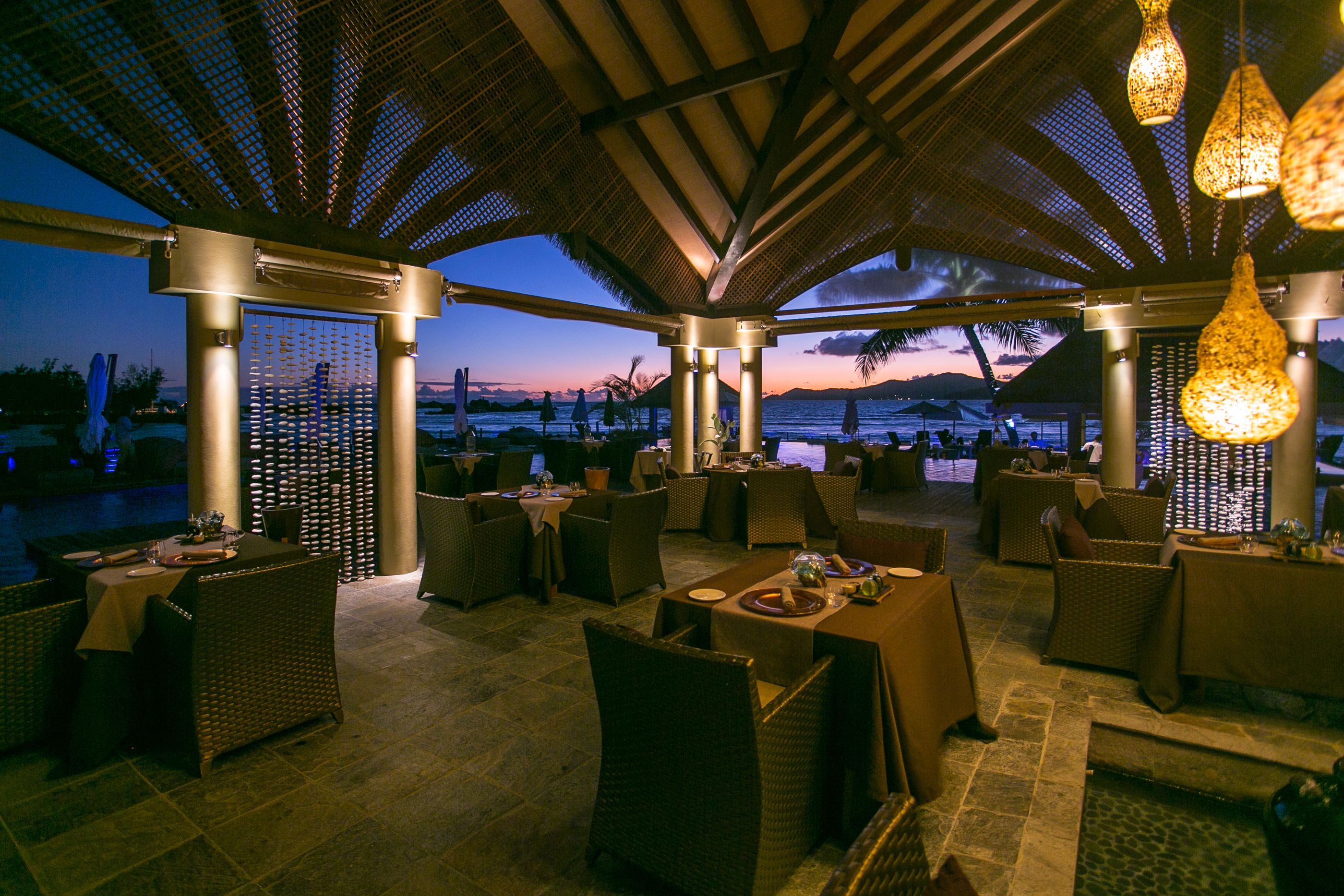 Un área de comedor al aire libre con muebles de mimbre y mesas dispuestas bajo un techo cubierto con luces colgantes.
