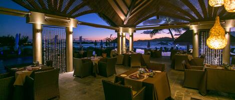 An outdoor dining area with wicker furniture and tables set up under a covered roof with hanging lights.