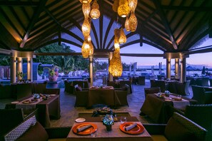 Un espacio de comedor al aire libre con mesas y sillas, luces decorativas colgantes y vistas al paisaje.