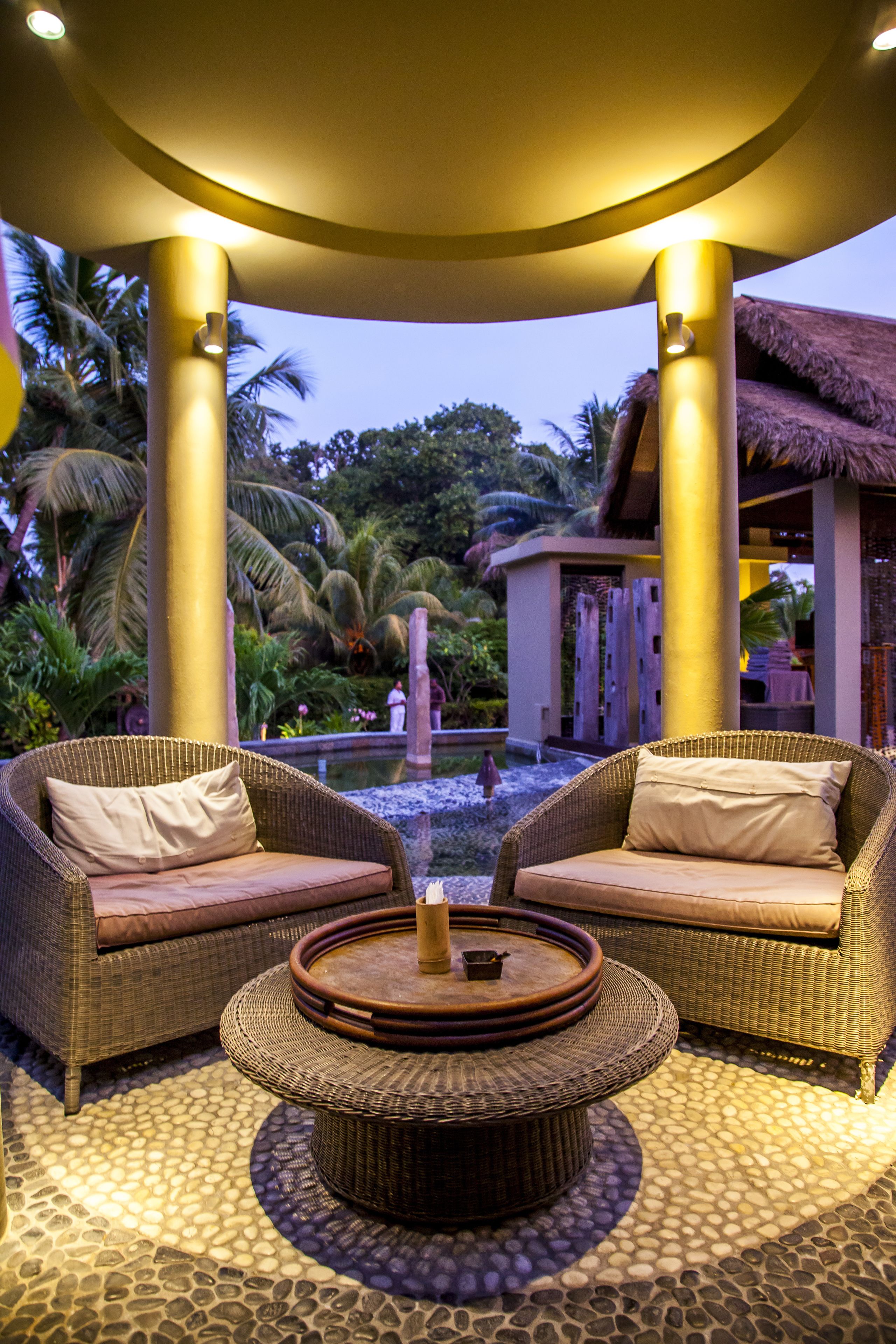 A circular seating area with wicker furniture and a central table, surrounded by a stone-tiled floor and palm trees in the background.