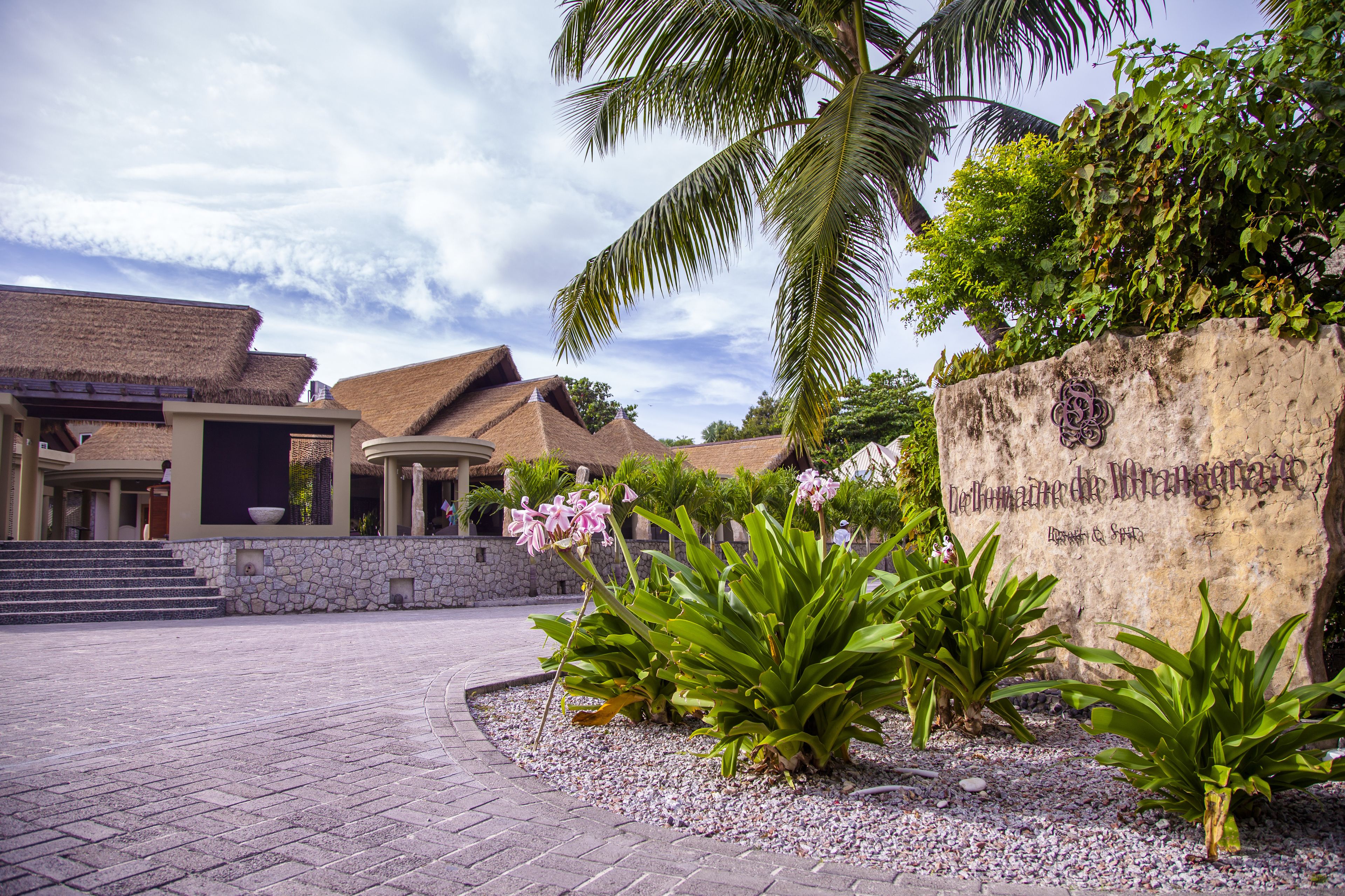A resort with thatched-roof buildings, a stone wall with a carved inscription, and tropical plants.