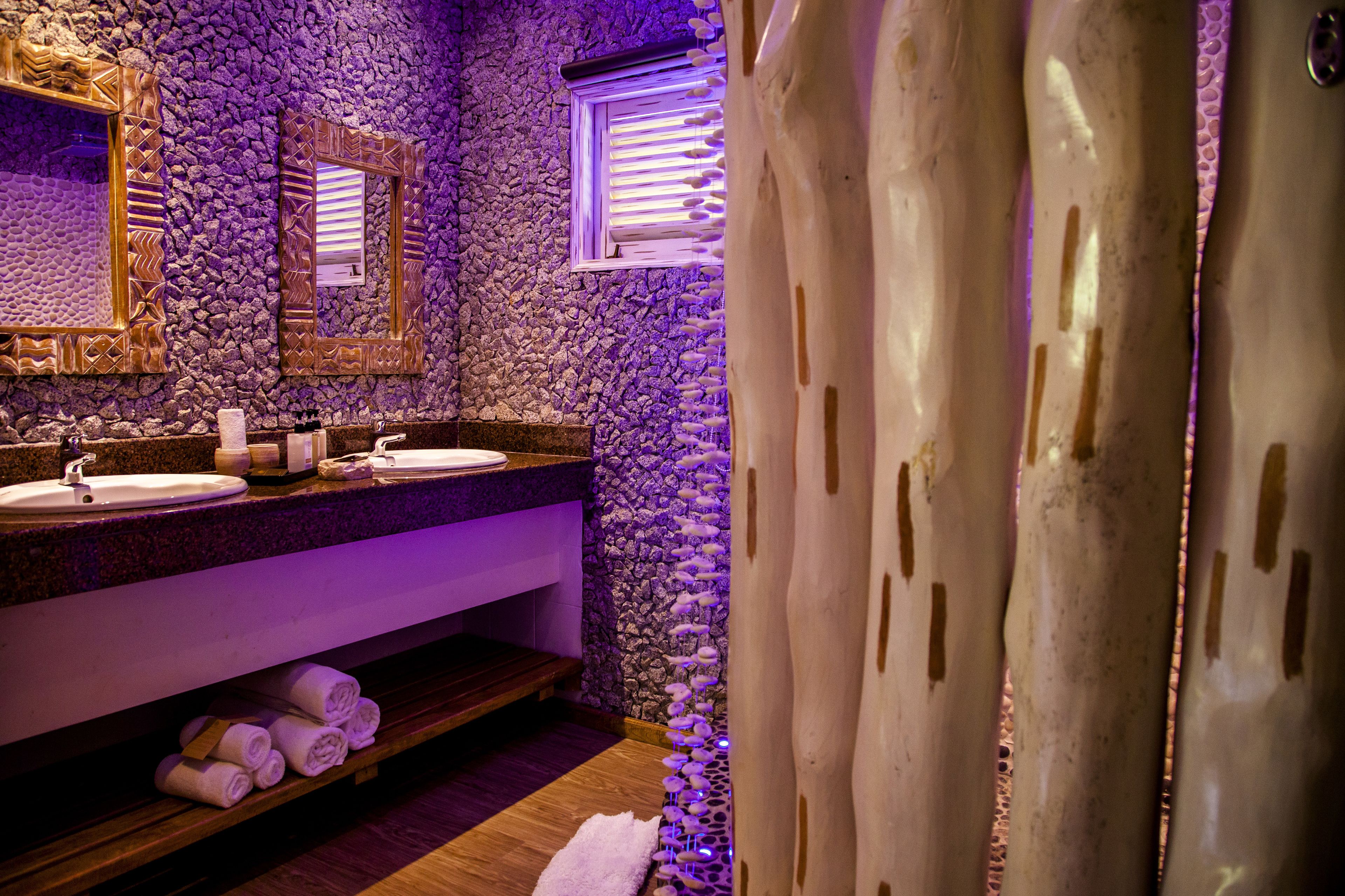 A bathroom with a stone wall, two sinks, a mirror, and a window with blinds.