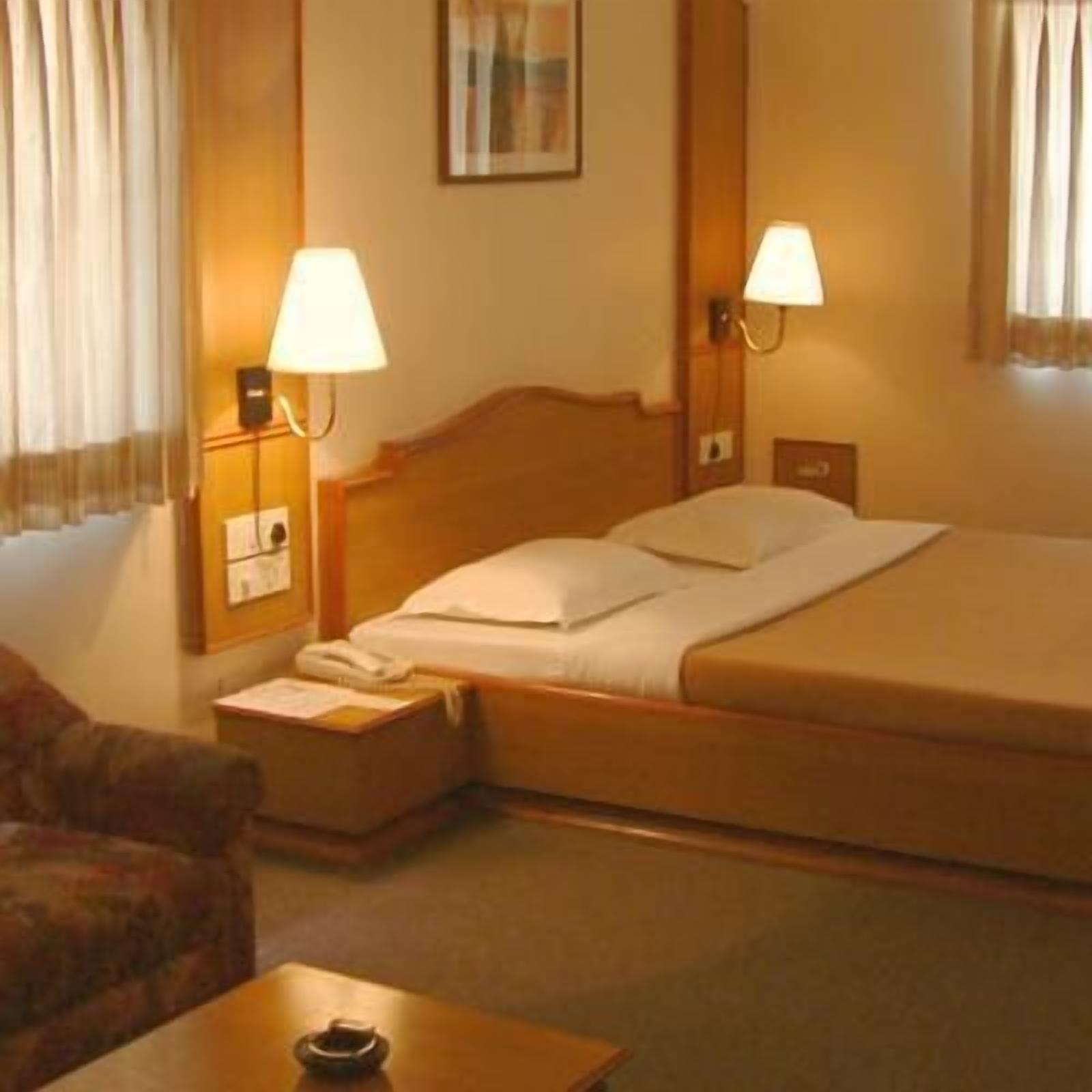 Habitación de hotel con una cama individual, dos lámparas de noche, una mesita de noche con un teléfono, una pequeña mesa con una taza y una silla.
