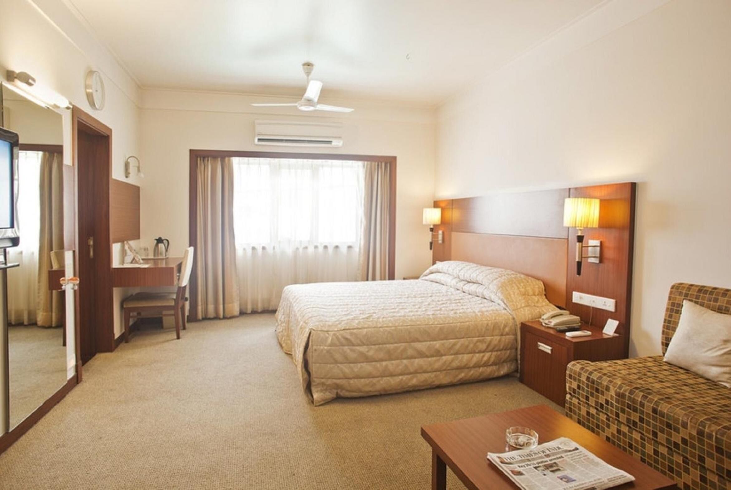 A hotel room with a bed, a desk, a chair, a small table, a nightstand, a lamp, and a television.