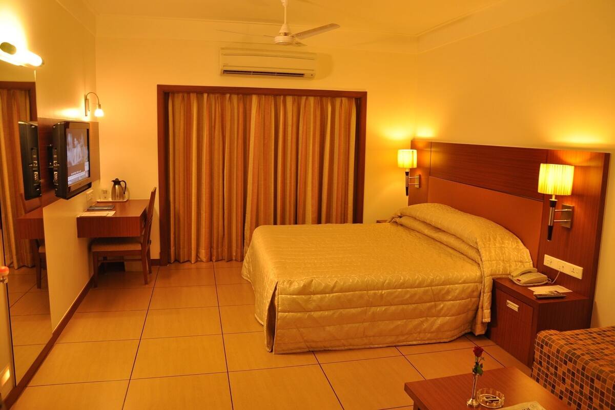 A hotel room with a bed, a desk with a television, a bedside table with a lamp, and a window with curtains.
