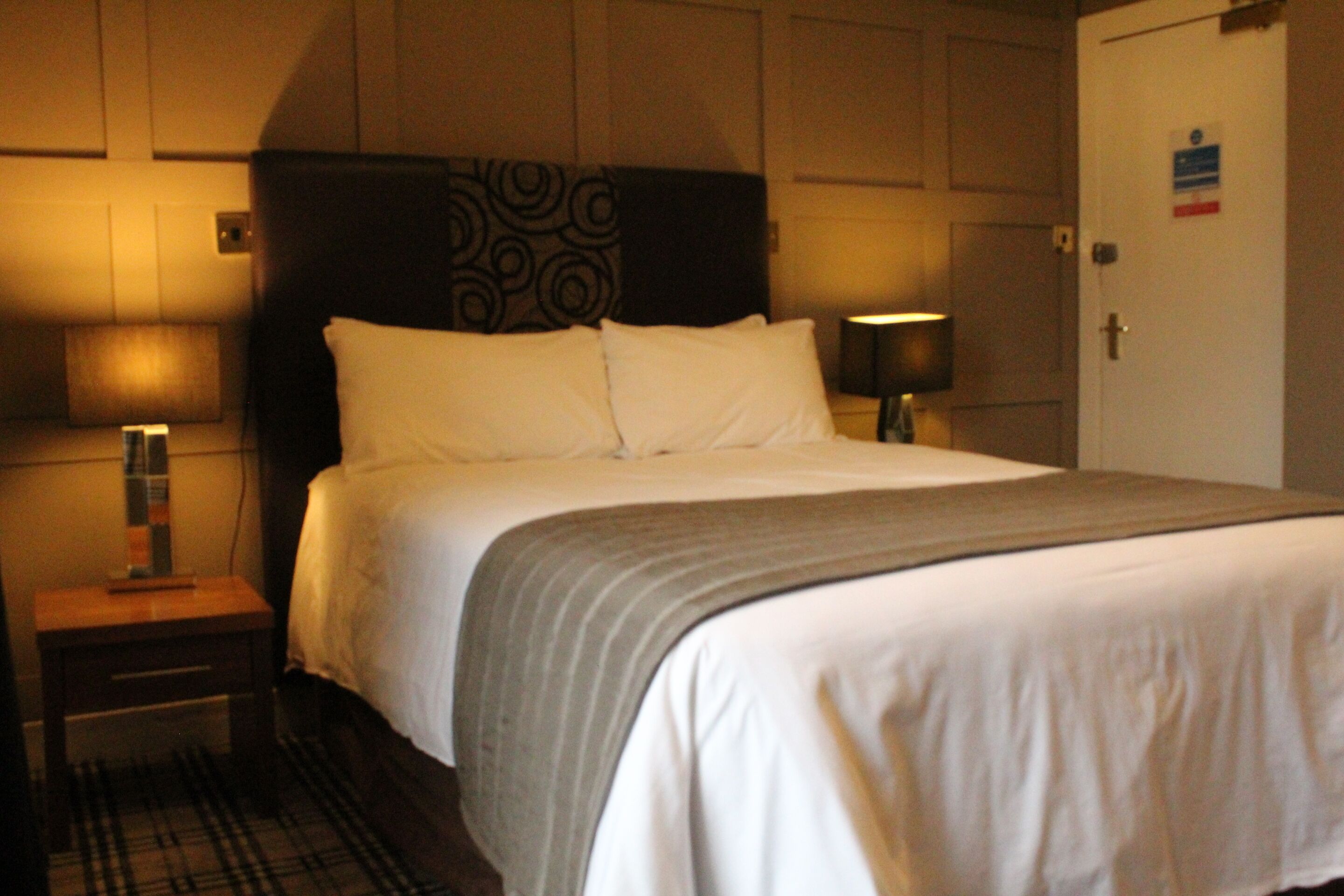 A hotel room with a large bed, a bedside table with a lamp, and a door with a sign.