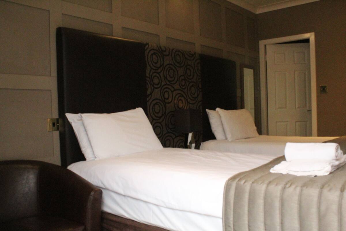 Een hotelkamer met twee bedden, een donker hoofdbord, een gordijn met patroon en een witte deur.