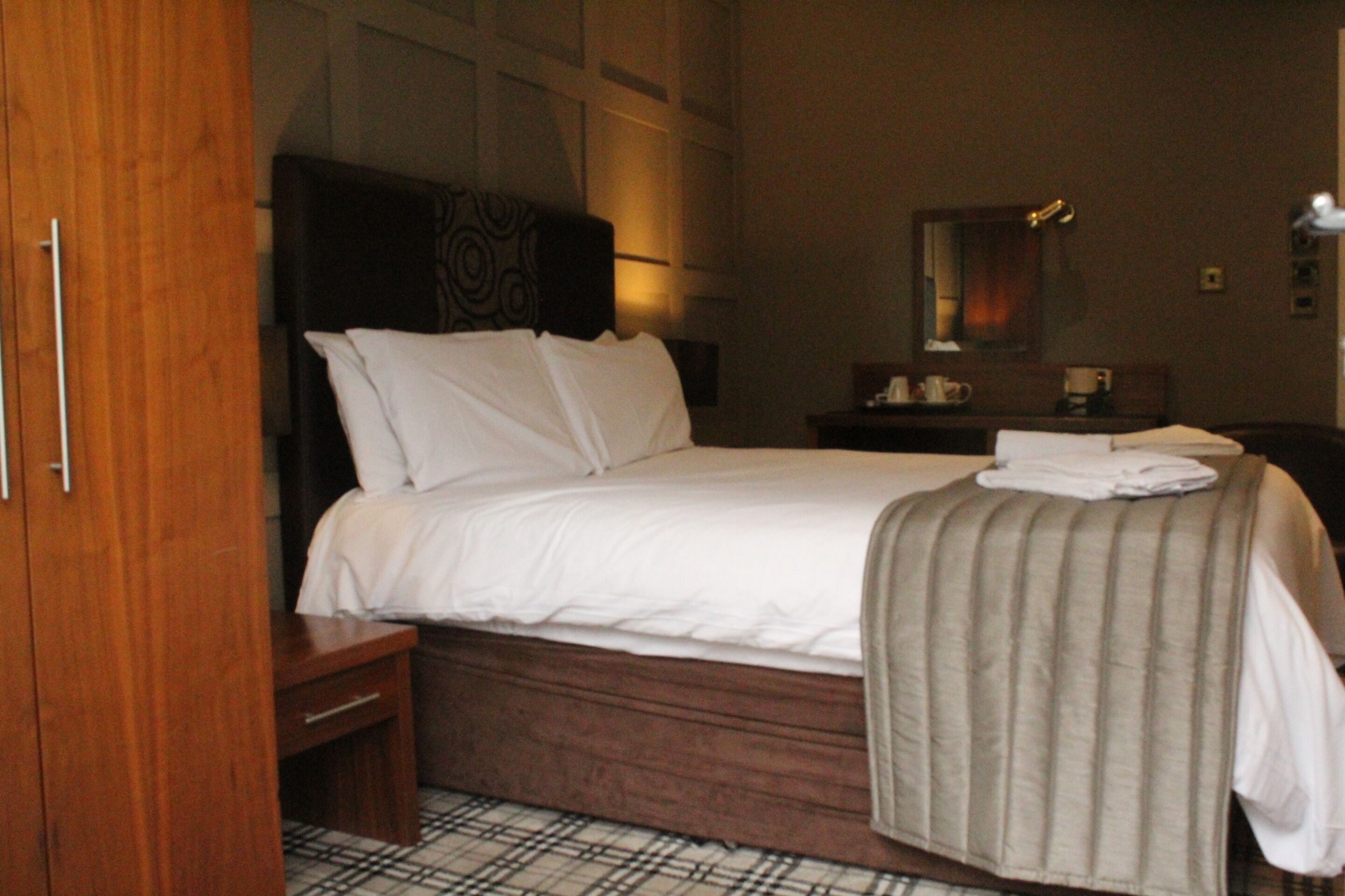 Habitación de hotel con una cama grande, una mesita de noche de madera, una mesilla, un espejo y una pequeña mesa con una taza y una caja de pañuelos.