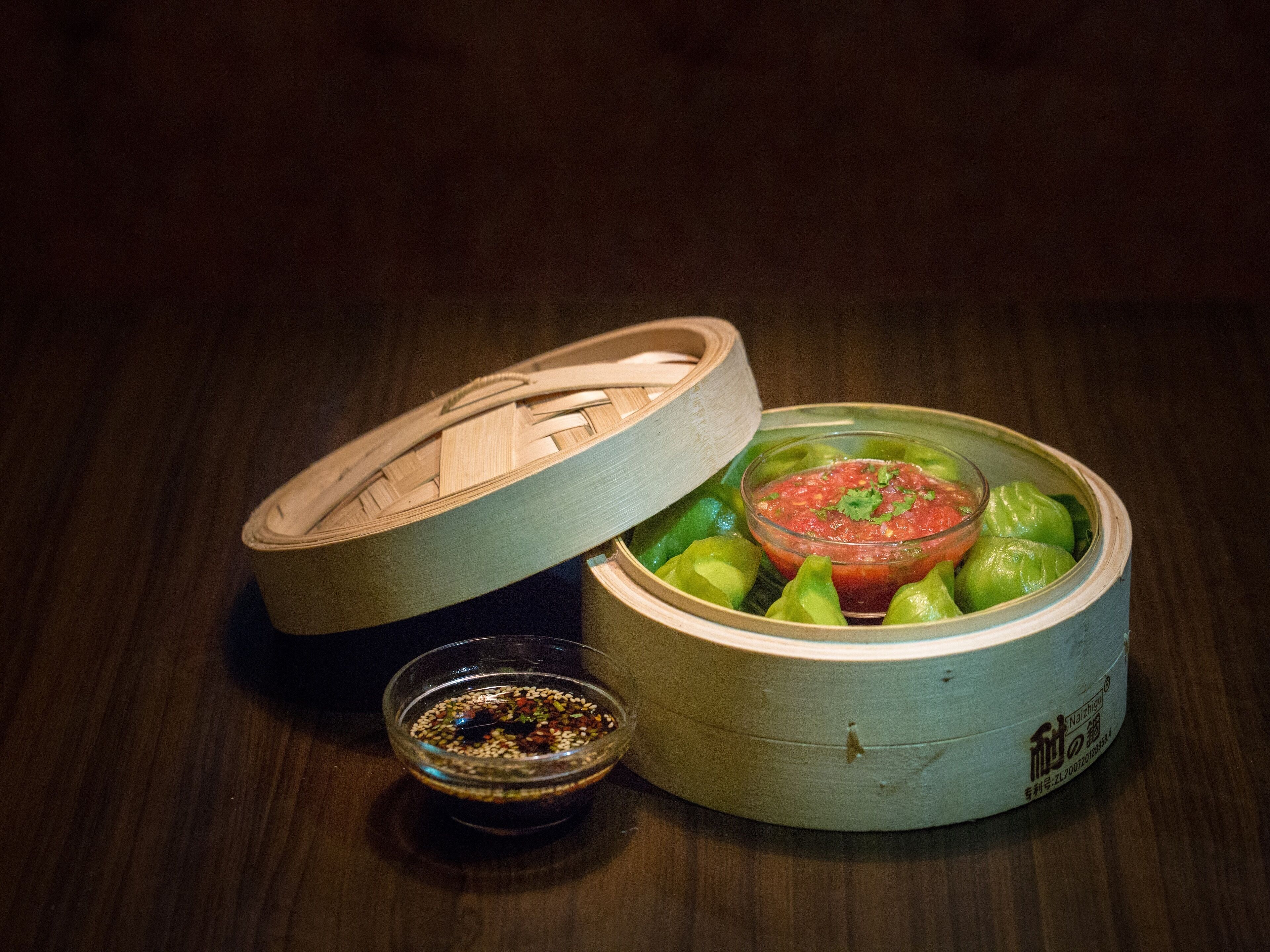 A bamboo steamer with green vegetables and a bowl of sauce.