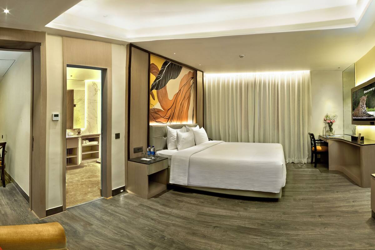 A modern hotel room with a large bed, a desk with a flat-screen TV, a seating area with a chair, and a bathroom visible through an open door.