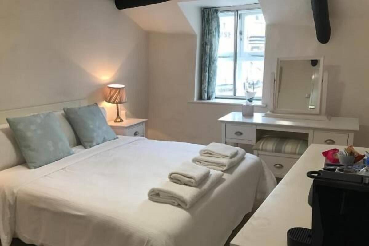 Ein ordentlich bezogenes Bett mit Kissen, eine Nachttischlampe, ein Schreibtisch mit Spiegel und ein Fenster mit Vorhängen.