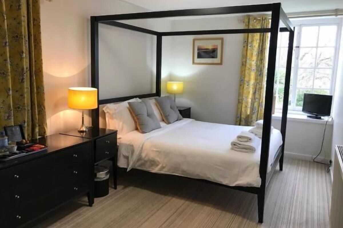 Ein Bett mit Baldachin, ausgestattet mit weißer Bettdecke und Kissen, ein Nachttisch mit Lampe und ein Fenster mit Vorhängen.