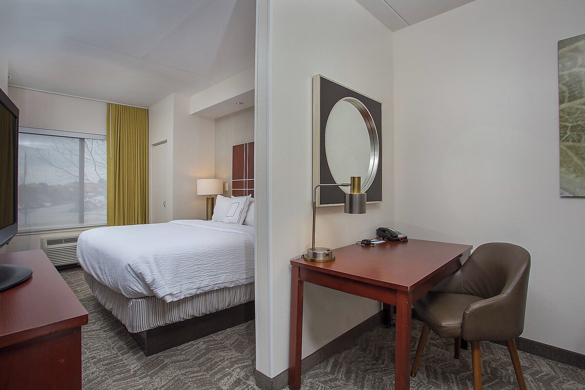 A hotel room with a bed, a desk, a chair, a desk lamp, a telephone, and a large mirror on the wall.