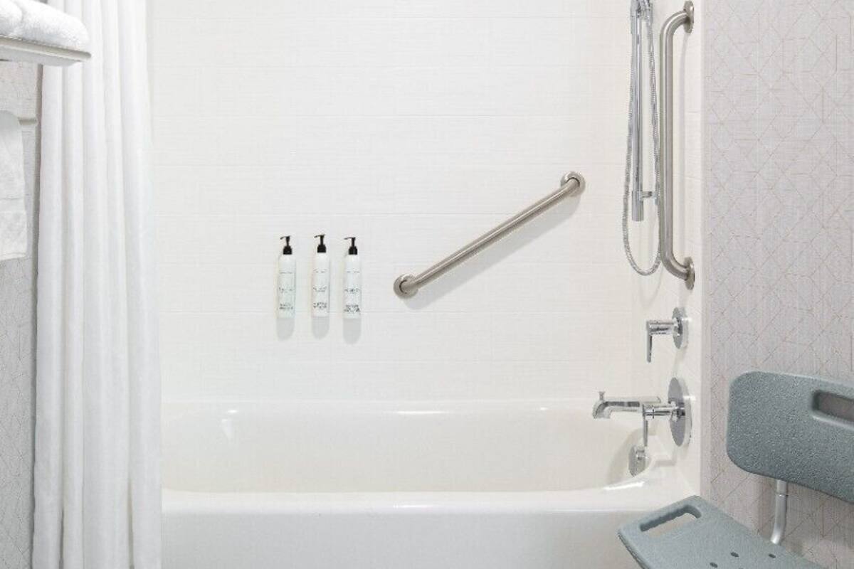 A white bathtub with a showerhead, grab bars, and a handheld shower attachment. There is a towel rack and a bench with a hole in the seat.