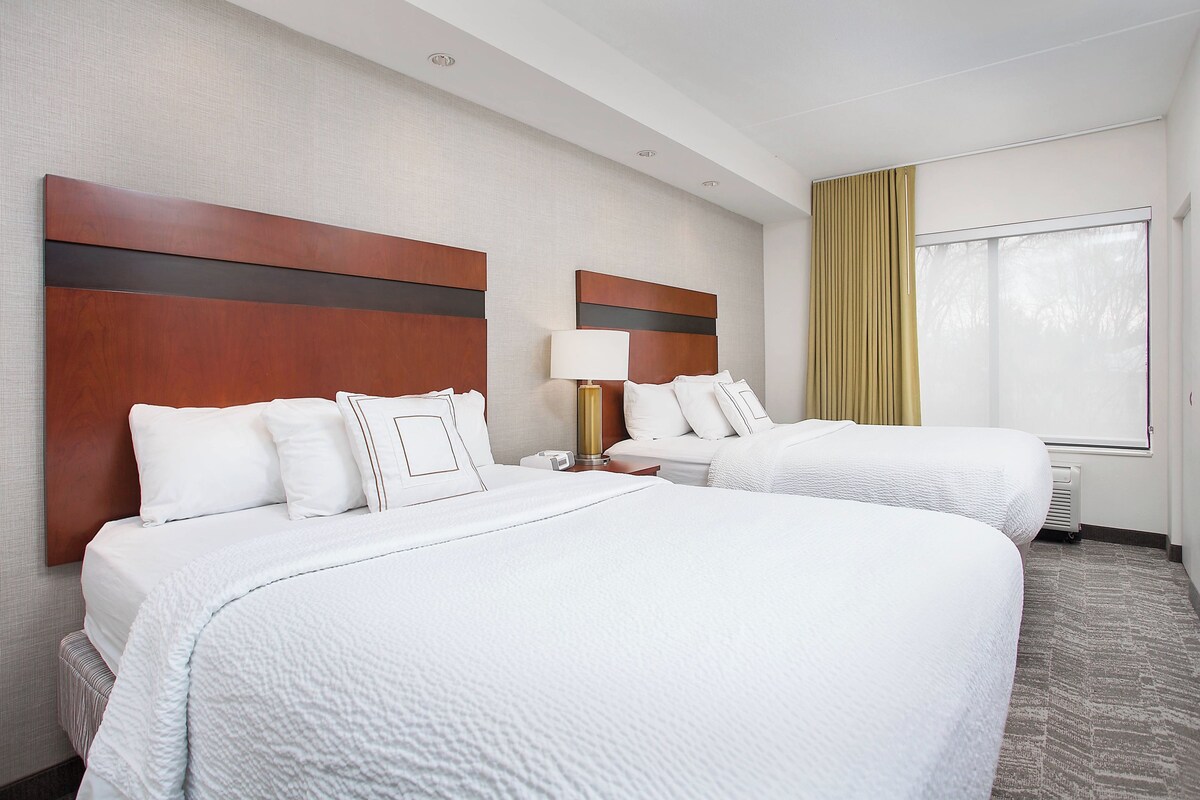A hotel room with two beds, a large window with curtains, and a wall-mounted lamp.