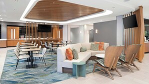 A modern hotel lobby with a curved wooden ceiling, a seating area with tables and chairs, a sofa with cushions, and a flat-screen TV mounted on the wall.