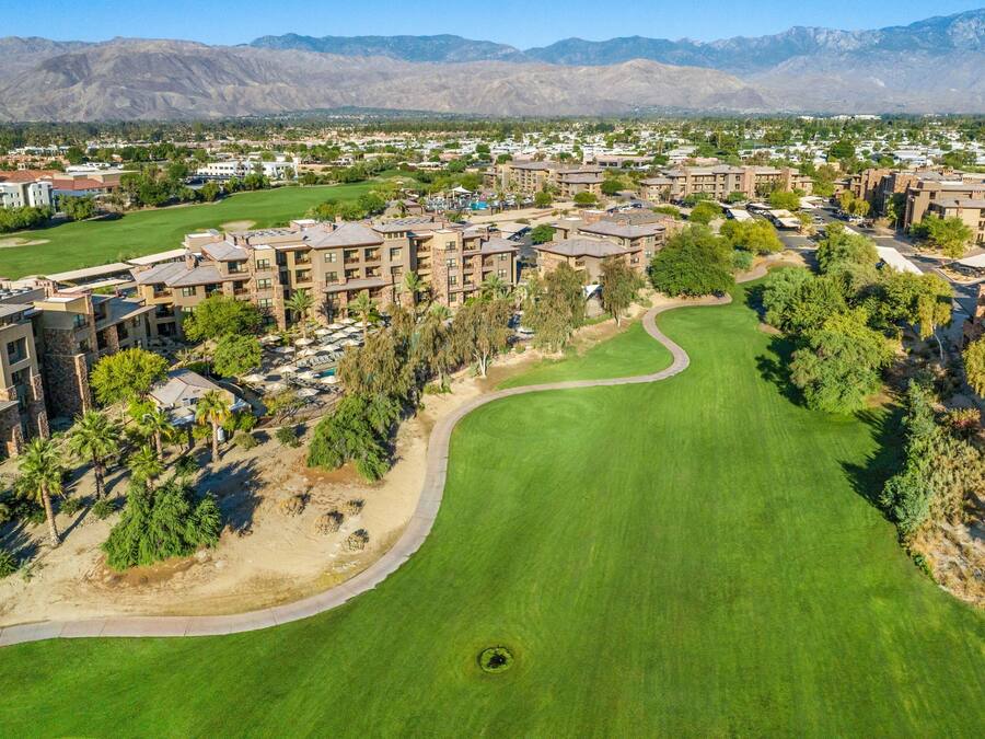 An aerial view of a golf course with a clubhouse, surrounded by residential buildings and mountains in the background.