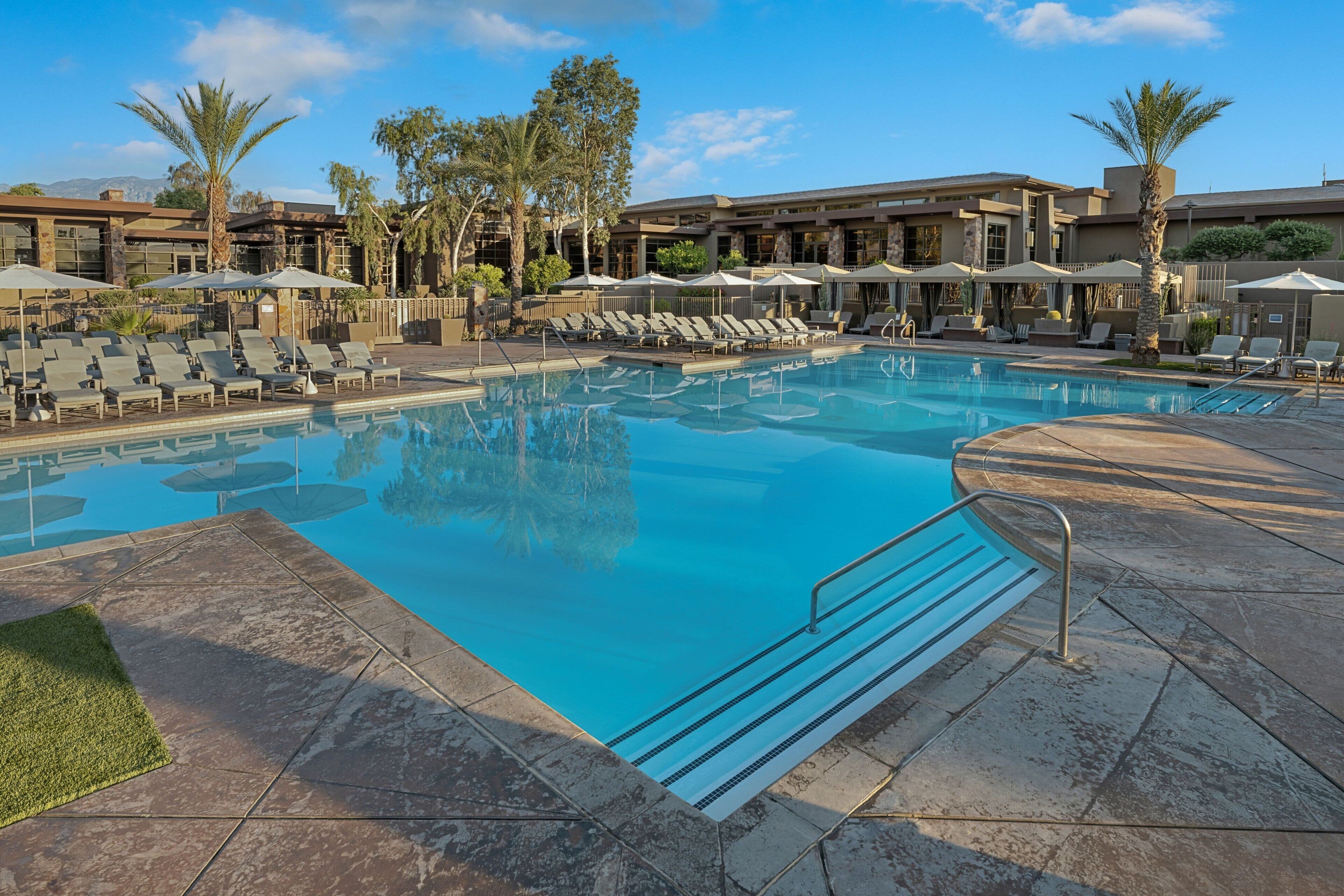 A swimming pool with lounge chairs and umbrellas, surrounded by palm trees and a building with large windows.