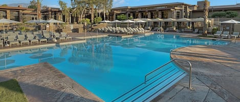 A swimming pool with lounge chairs and umbrellas, surrounded by palm trees and a building with large windows.