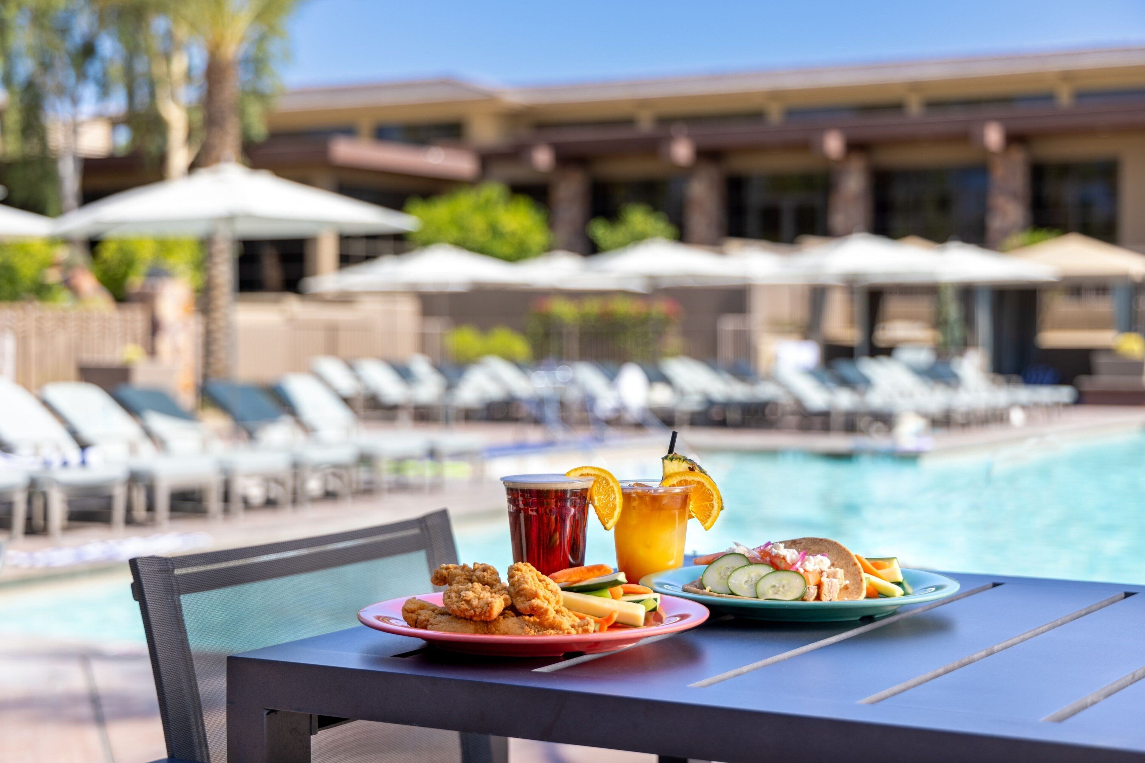 A dining table with plates of food and drinks by a poolside, with lounge chairs and umbrellas in the background.