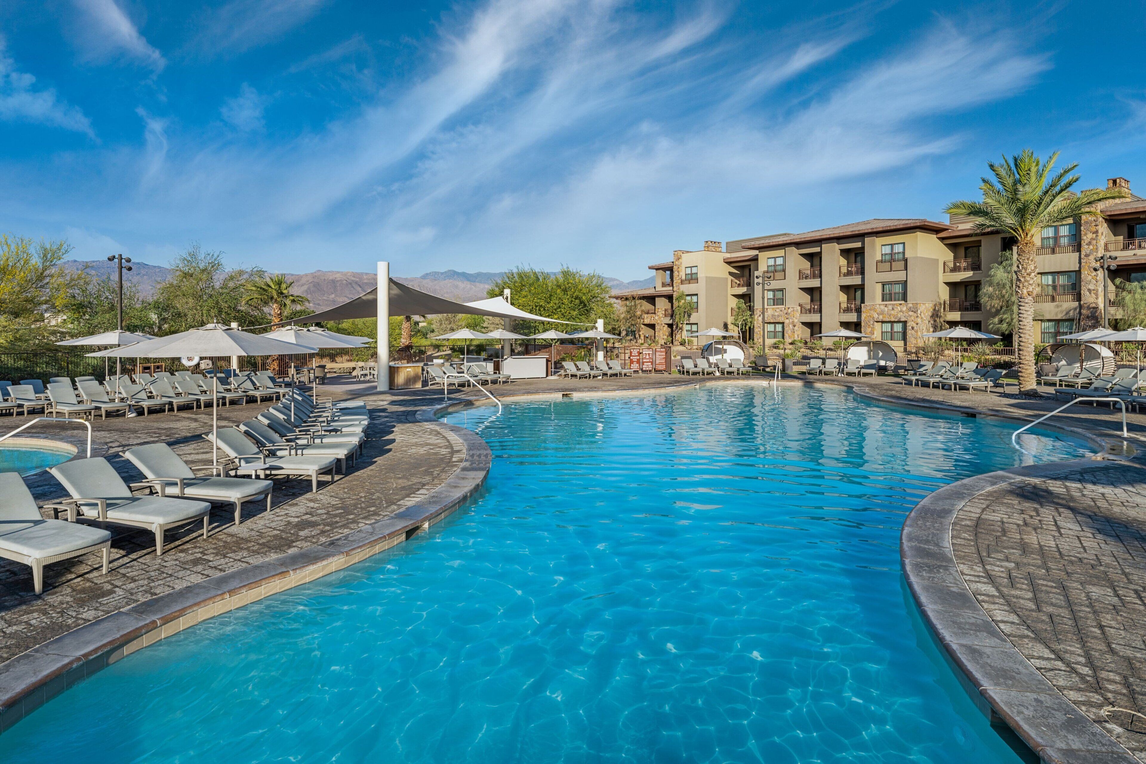 A pool area with lounge chairs and umbrellas, surrounded by a building with multiple balconies and a mountain view in the background.