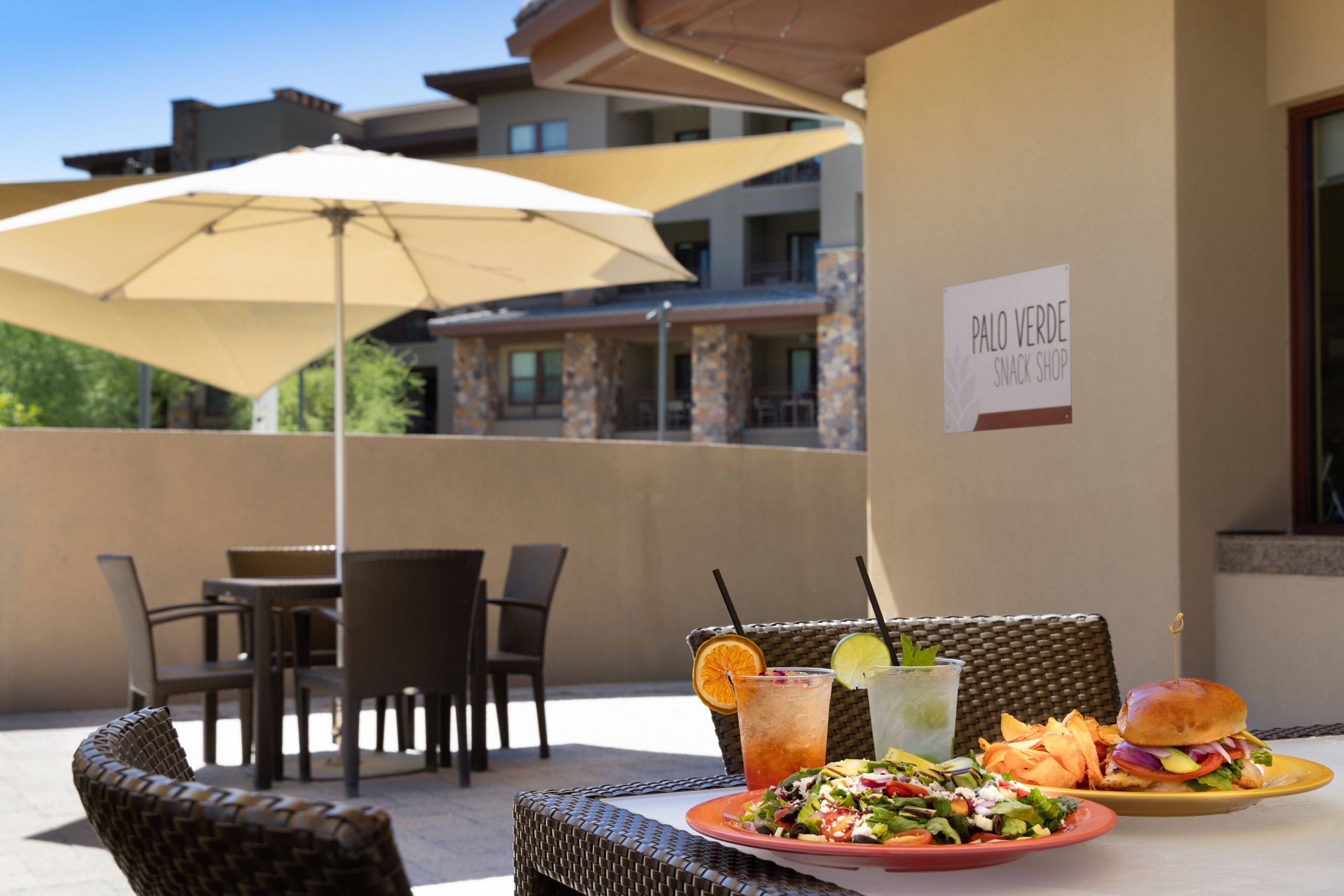 An outdoor dining area with wicker chairs and a table, a large umbrella, and a sign for Palo Verde Snack Shop.