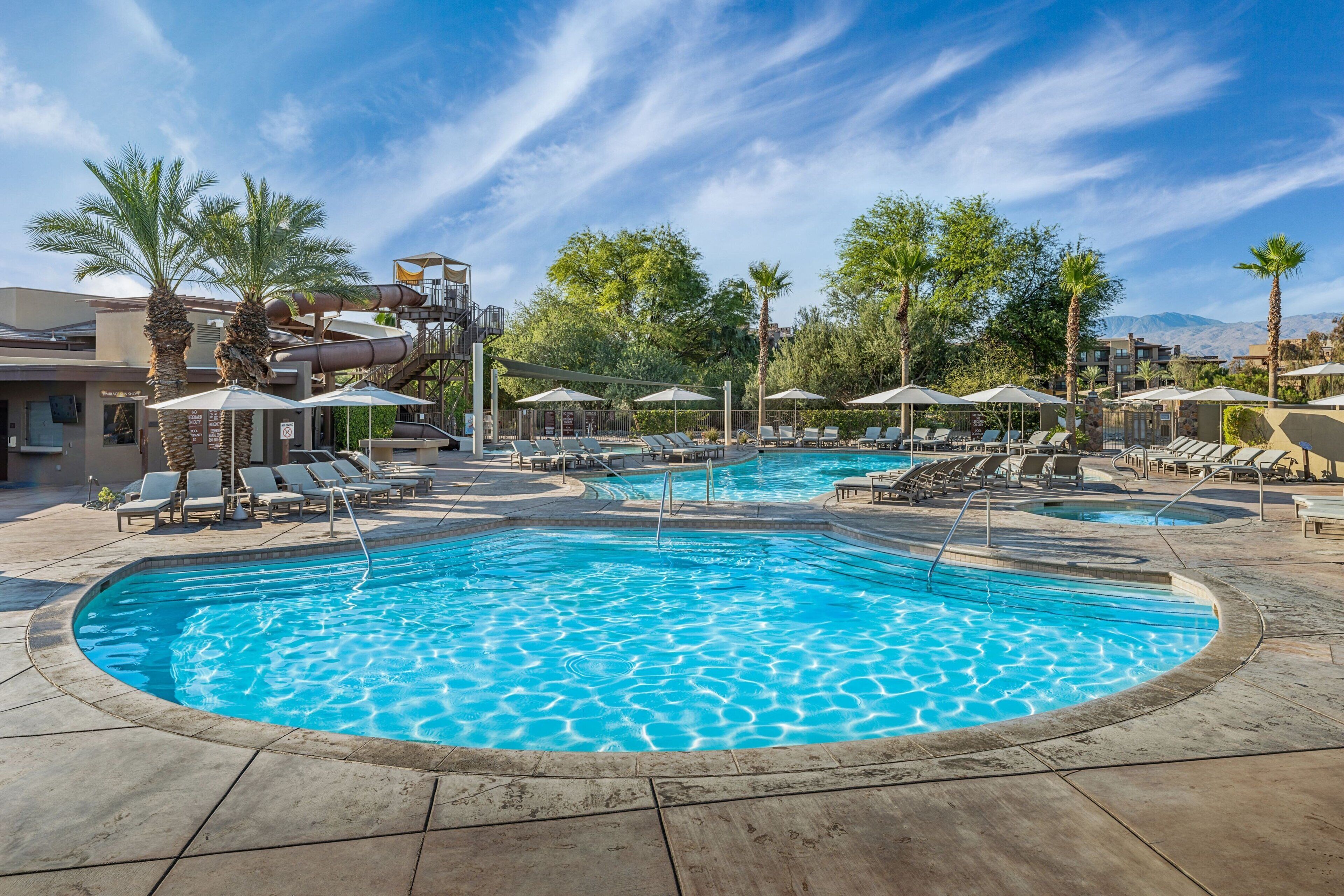 A pool area with multiple pools, lounge chairs, umbrellas, and palm trees.