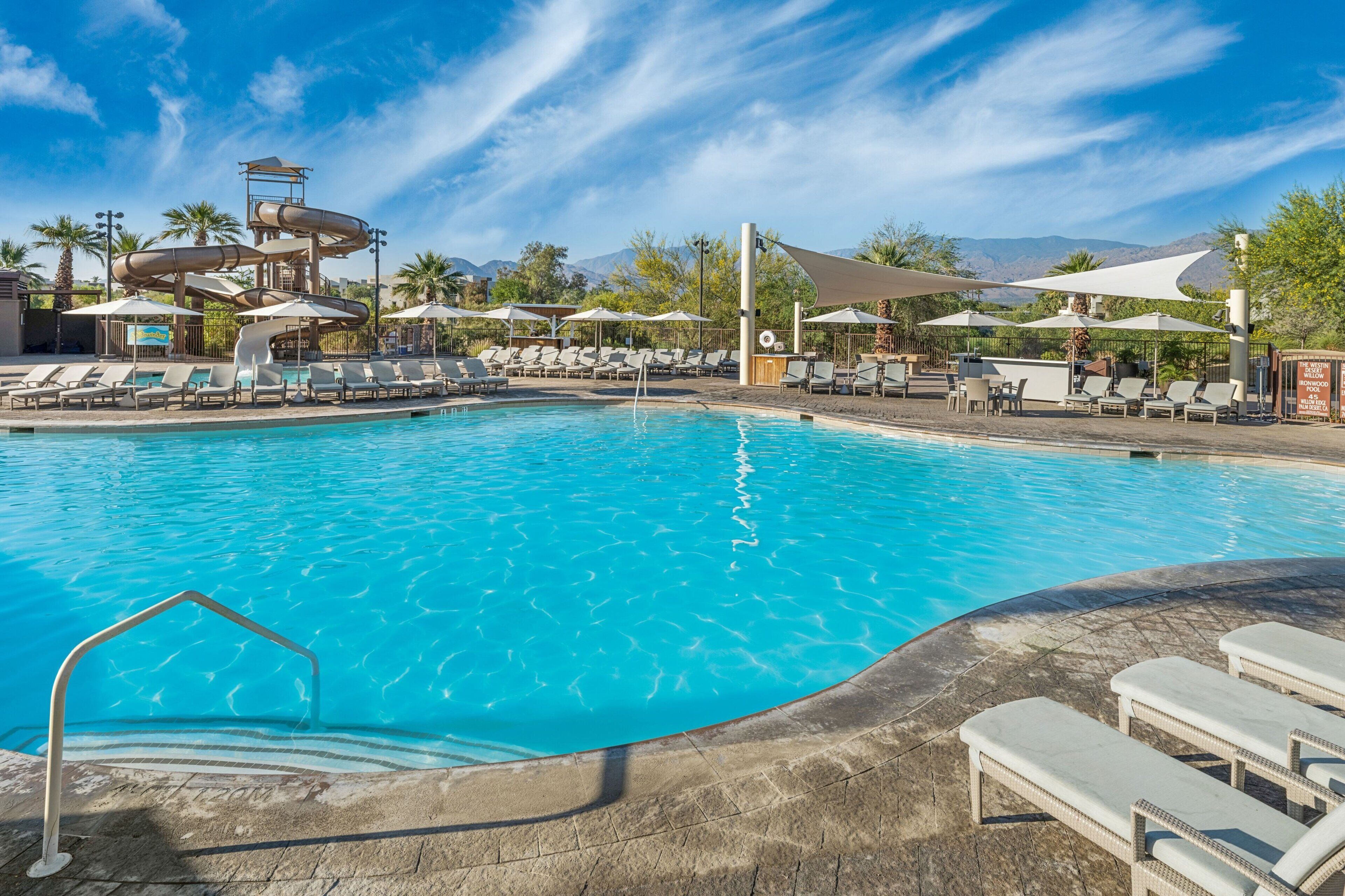 A swimming pool with lounge chairs and umbrellas, a water slide, and mountains in the background.