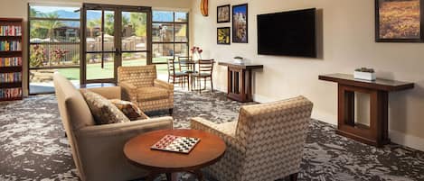 A spacious living room with a large window, a bookshelf, a flat-screen TV, and a chessboard on a coffee table.